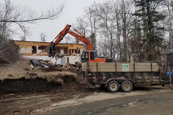 Excavator mid demolition of an old house with pickup truck parked in front